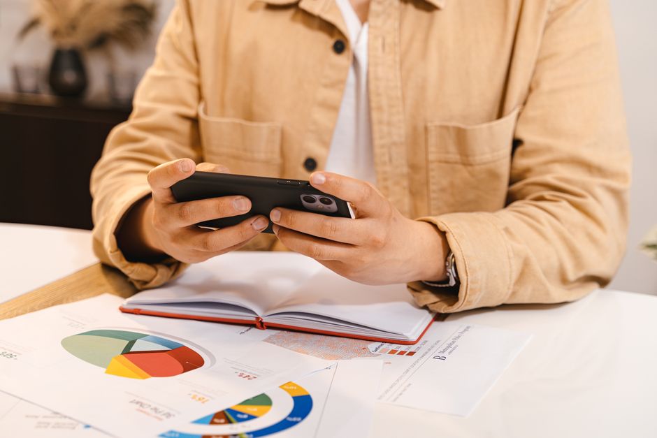 Person in jacket using smartphone, reviewing charts and data at a workspace.